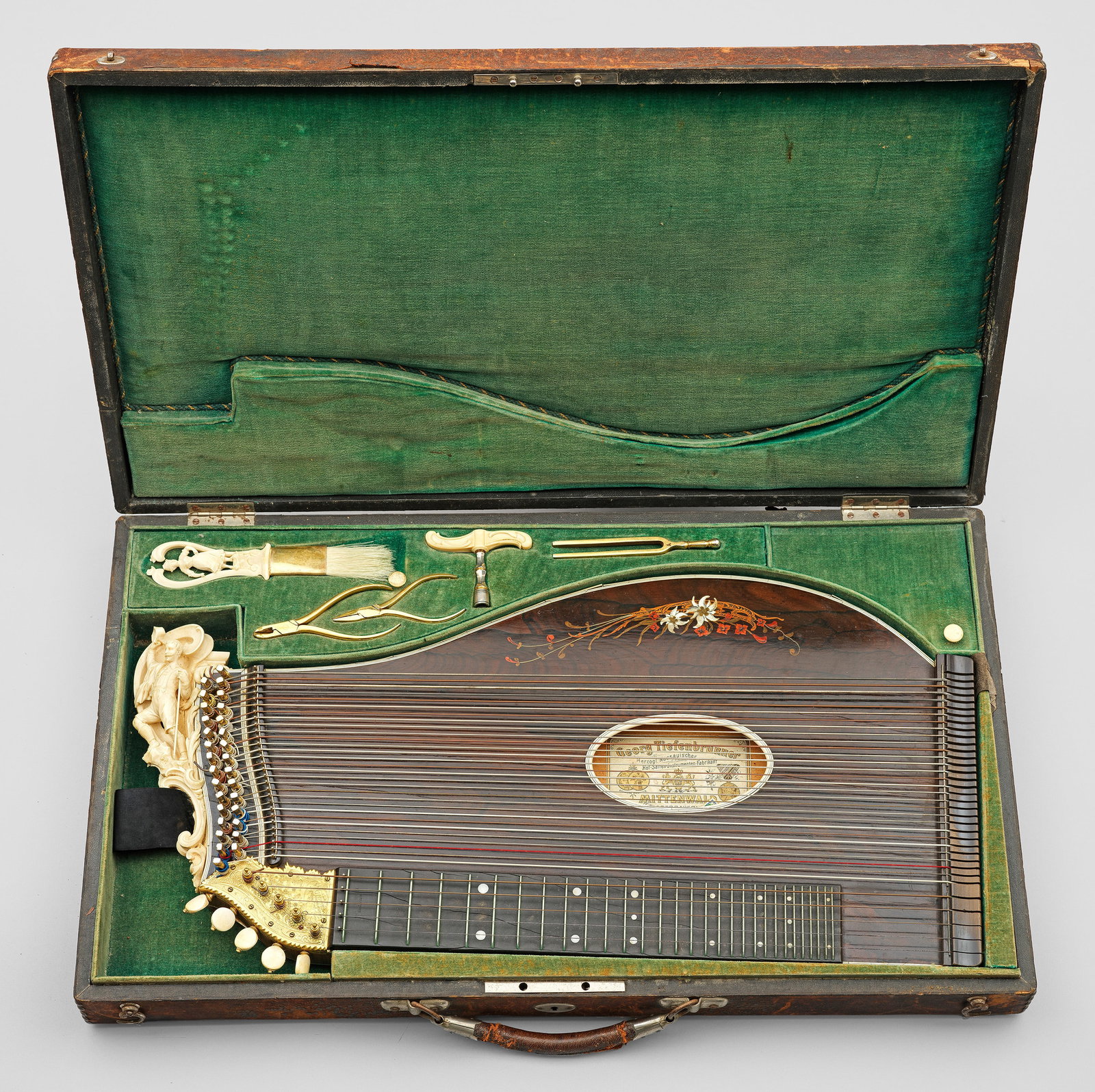 Konzert-Zither von Georg Tiefenbrunner: Holz, furniert und Elfenbein, reliefplastisch geschnitzt. 5 Griff- und 29 Freisaiten. Korpus mit eingelegter, polychromer Blumendarstellung. Seitliche, vollplastische Elfenbeinschnitzerei in Form eine
