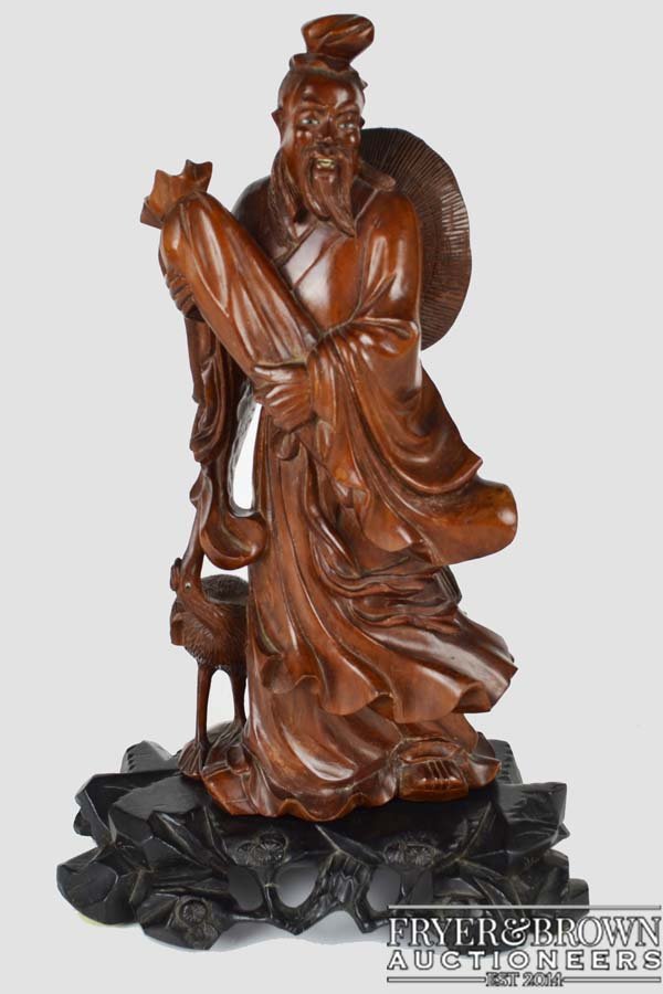 A Chinese carved wood figure of an Immortal standing: A Chinese carved wood figure of an Immortal standing holding a long scroll bag with crane to one side, inlaid glass eyes with straw hat on his back, on a black hardwood prunus and rock work base, 36cm