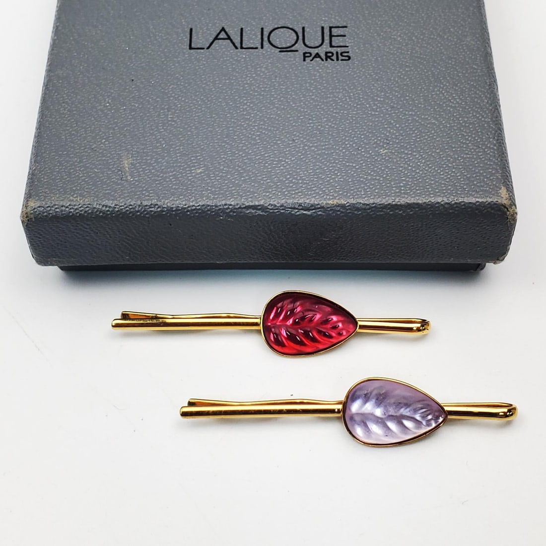 2 Lalique France Heliconia Glass Leaf Hair Pins: Lot of 2 Lalique France "Heliconia" Glass Leaf Hair Pins. In Original Box w/ Original Bag. Condition: Generally Very Good. Dimensions: L-2" Each. Provenance: From a Long Island, NY Home. Please note t