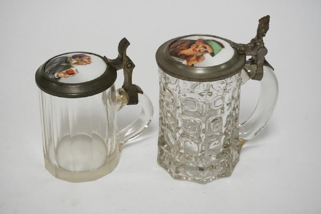 PAIR OF GLASS STEINS WITH PAINTED PORCELAIN LIDS: PAIR OF GLASS STEINS WITH PAINTED PORCELAIN LIDS DEPICTING A PENSIVE MAN AND A WOMAN DRINKING WINE. TALLEST IS 6 3/4 INCHES.