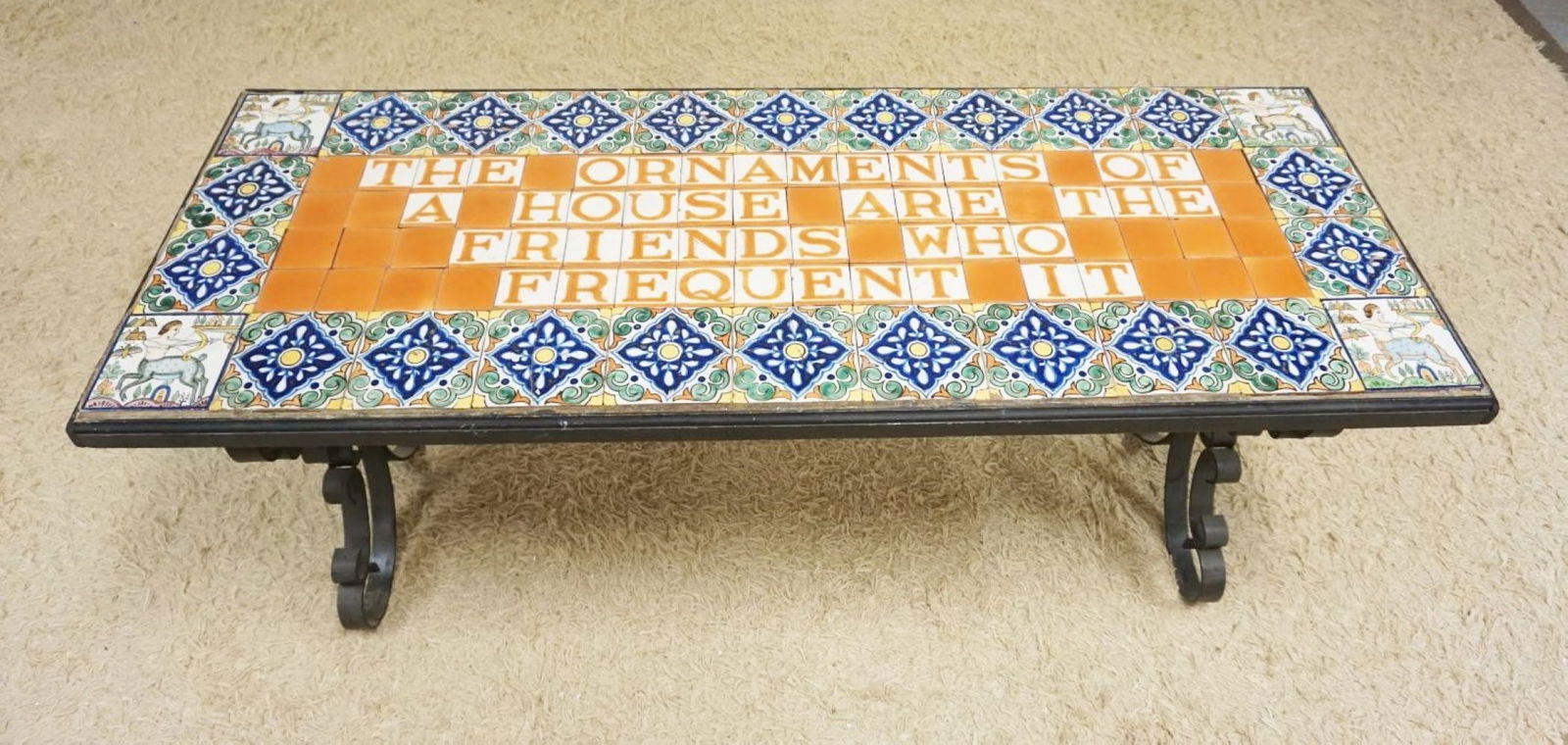 TILE TOP TABLE: TILE TOP TABLE ON SCROLLED IRON BASE, THE ORNAMENTS OF A HOUSE ARE THE FRIENDS WHO FREQUENT IT, SOME TILE DAMAGE, APPROXIMATELY 24 IN X 63 IN X 26 IN H