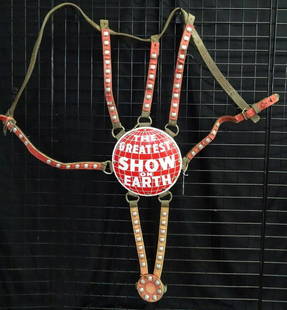RINGLING BROS. BARNUM BAILEY HEAD DRESS: ORIGINAL RBBB GREATEST SHOW ON EARTH ELEPHANT HEAD DRESS. INCLUDES PORCELAIN MEDALION MOUNTED ON LEATHER BACKING WITH CONCHO ADORNED LEATHER STRAPS. CRACKS IN THE STRAP COVERING. "ARSAN" TOOLED