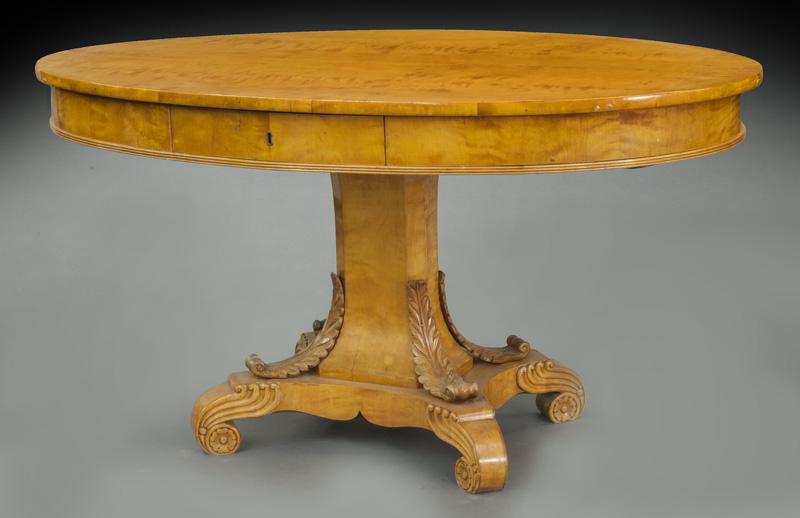 Biedermeier fruitwood 2-drawer library table: Biedermeier fruitwood 2-drawer library table the oval top standing on a shaped rectangular pedestal, on a platform base with applied acanthus carvings and carved scrolled feet. 29.5"H x 57"W x 36.25"D