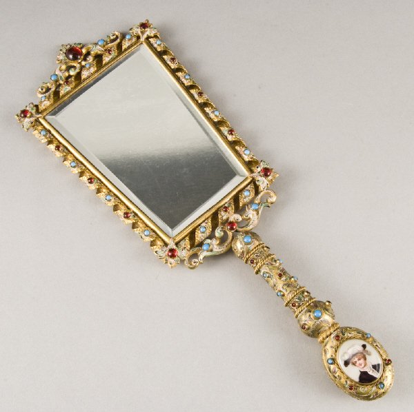 French gilt metal and enamel hand mirror: the edges and handle with polychrome enamels and inset cabochons of turquoise and ruby glass, the end of the handle inset with hand painted porcelain plaques. 13.75"H x 5"W, Circa - Late 19th C.