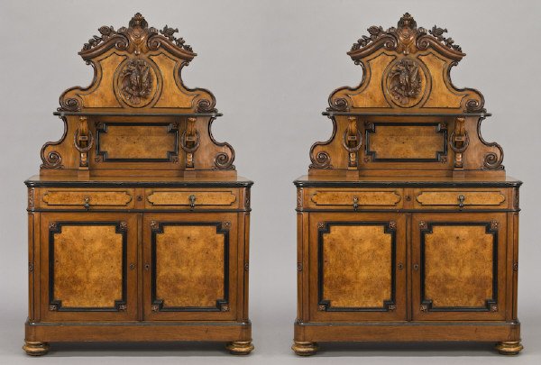 Pr. Victorian walnut hunt cabinets,: each with a highly carved and scrolled pediment centered by a trophy, over a single shelf, the cabinet having two short drawers and a pair of paneled doors resting on bun feet. 84"H x 55"W x 21"D, Cir