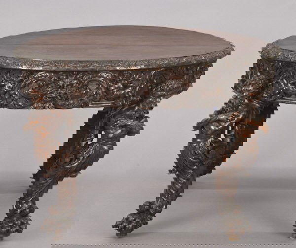 Italian Renaissance Revival walnut dining table: having a round carved top enclosing three leaves within a foliate carved edge above a deeply carved apron depicting cherubs interspersed with grotesque masks on a scroll foliate ground, raised on four