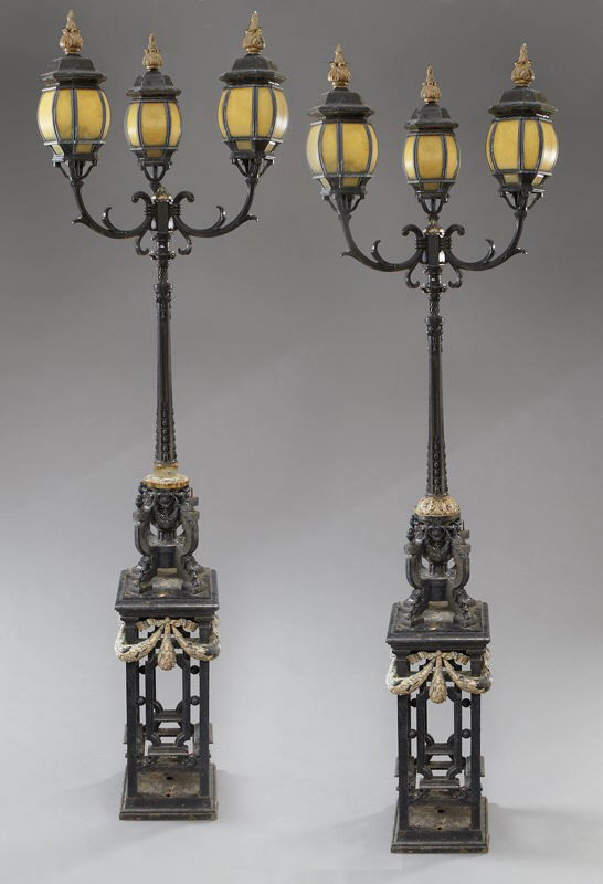Pr. wrought iron street lamps, each with three: Pr. wrought iron street lamps, each with three lanterns with glass panels raised on scrolled arms, resting on a pierced plinth base draped with laurel garland. 103" H x 13.5" Sq. at base, Circa - 20th