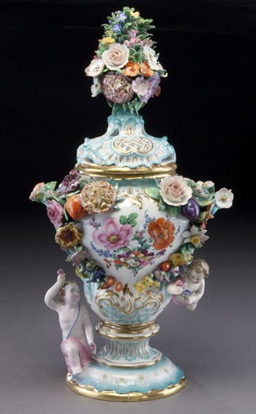 Meissen porcelain lidded vase with a floriform: finial and pierced lid; the handled body with heavy overall applied floral decoration as well as hand painted floral reserves, and two putti. Cross swords mark in blue under glaze. 17"H, Circa - Late