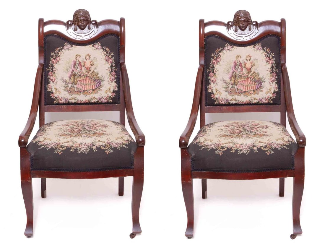 Pair of Victorian Needlepoint Armchairs: Wood armchairs with needlepoint upholstery. Carved faces at the top of each seatback. Measuring: H 40.75 x W 22.25 x D 20 inches Condition: Scratches and scuff to the wood in various areas 