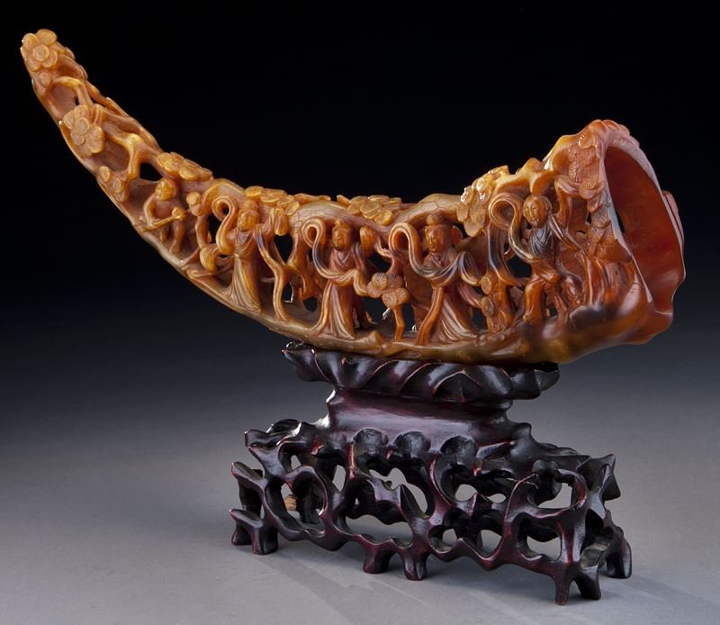 Chinese buffalo horn carving: Chinese buffalo horn carving depicting immortals and attendants in a pine grove, with exquisite details in high relief and openwork, presented on the original wood stand. 15"W, Circa - Early 20th C.