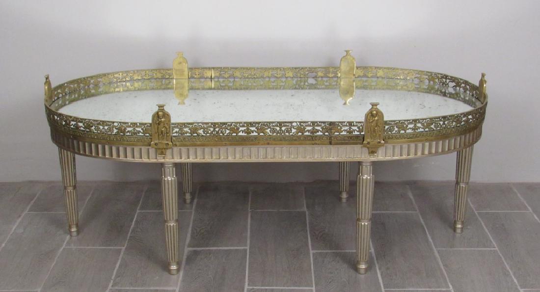 Neo-Classical Style Cocktail Table: Mirrored top with gallery having griffins and angel post. Silvered fluted base with six legs. 49"W 23"D 20"H.