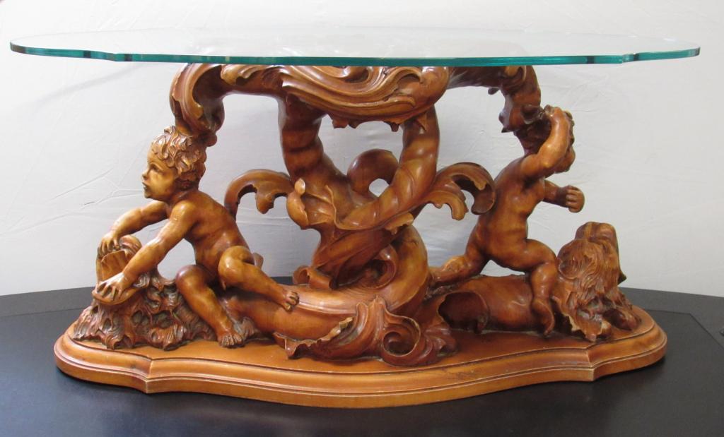 Carved Putti Figural Coffee Table: Carved wood 2 putti riding atop lion head sea serpents on a carved wave. Turtle shaped glass top, beveled wood shaped base. Base 43" X 18" - Glass 44" X 22"