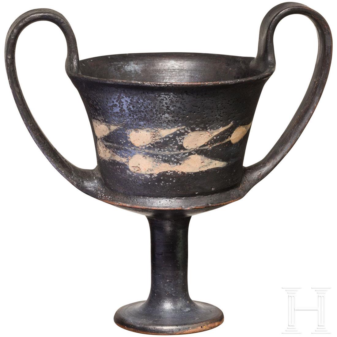A Boeotian black glazed kantharos, Greece, 5th century: A medium size kantharos with a deep bowl and tall pedestal foot with twin upward sweeping handles. It is decorated with white leaves. One handle broken and restored, otherwise intact. Height 17 cm. Pr