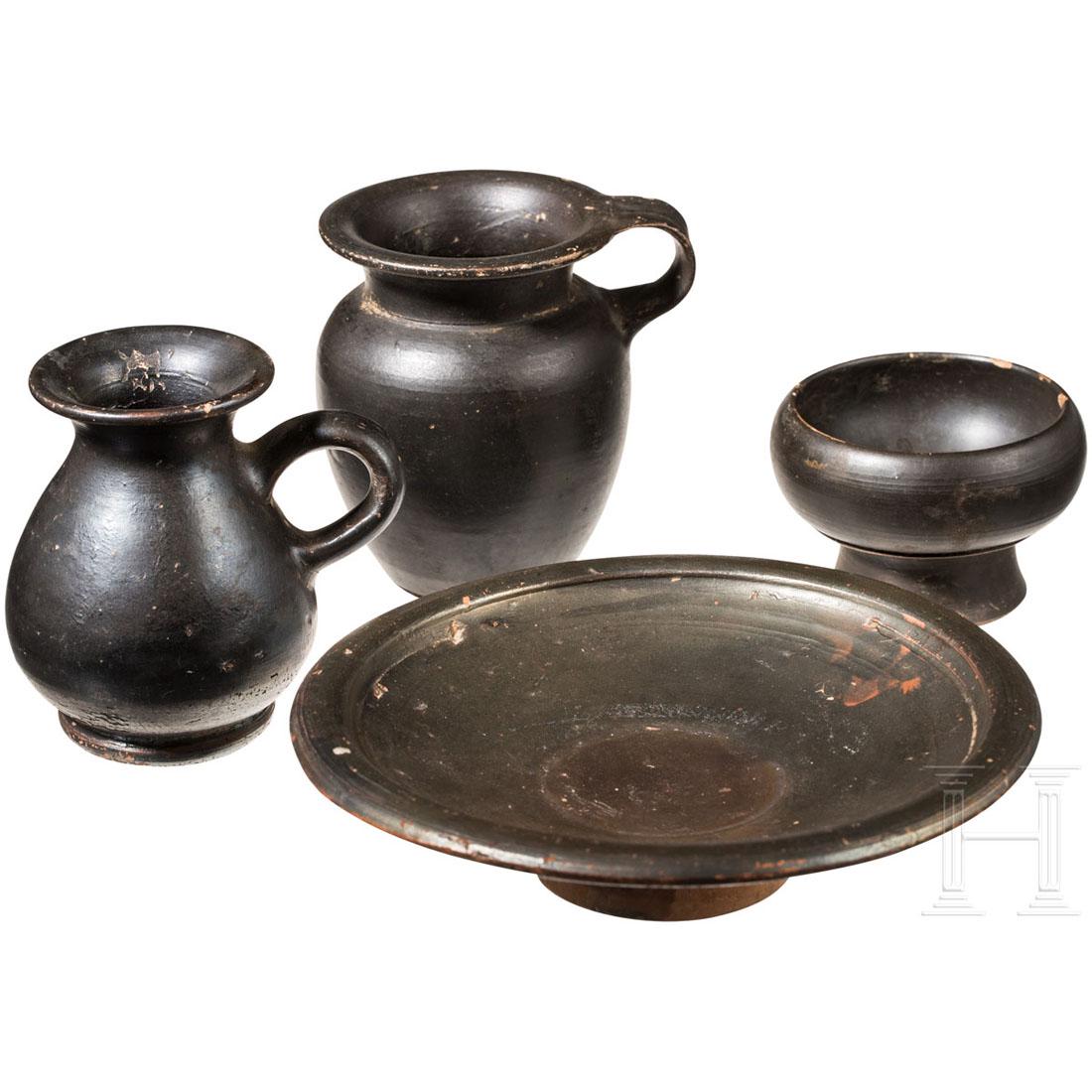 Four black glazed miniature vessels, Italy, 4th - 3rd: A flat bowl. An "N" engraved in western Greek script on the underside. Diam. 15 cm. A small drinking bowl on a high foot. Diam. 6 cm. And two small jugs. H. 8 cm and 8.5 cm. Vessels partially minimall