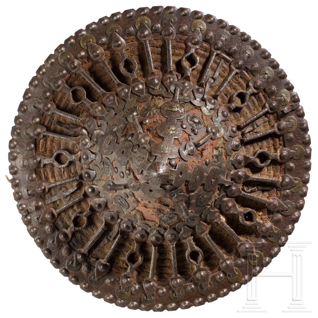 An iron-fitted Caucasian fist shield, circa 1900: GewÃ¶lbter Korpus aus (mit roter Wolle umwickeltem) Weidengeflecht. Die AuÃŸenseite eisenbeschlagen mit nietenbesetztem, konischem, spitz zulaufendem Buckel und durch zahlreiche Spangen verbundene