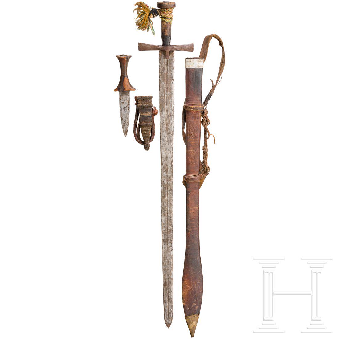 A kaskara and an arm dagger, North Africa, circa 1900: Kaskara mit langer, zweischneidiger Klinge mit beidseitig gestempelten Halbmondmarken, eiserner Parierstange und textilumflochtenem Holzgriff. Schauseitig punzierte Lederscheide mit Mund- und Ortbesch