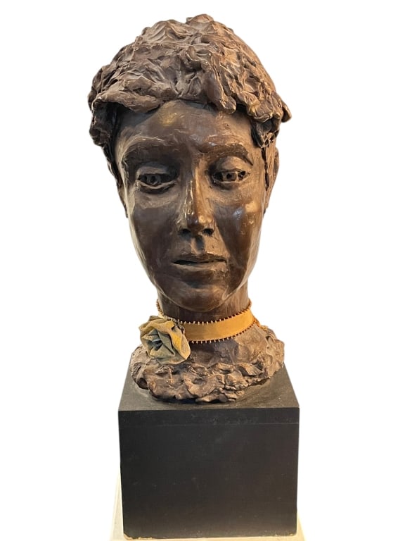 BRONZE-TONE FEMALE BUST SCULPTURE ON PEDESTAL BASE: DECORATIVE BRONZE-TONE FEMALE HEAD BUST SCULPTURE WITH RIBBON COLLAR Decorative bronze-tone female bust sculpture featuring a serene young woman’s face with textured hair and expressive hand-finishe