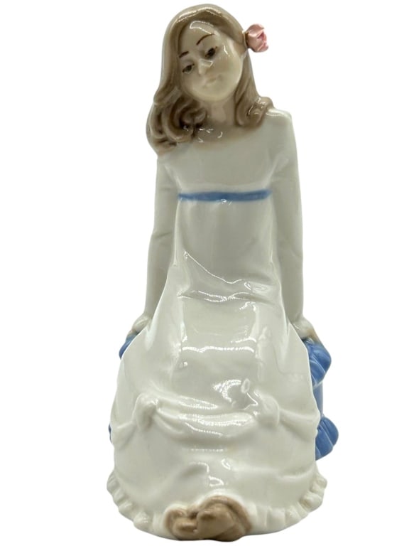 NADAL PORCELAIN GIRL FIGURINE 6": Sweet glazed porcelain figurine depicting a seated young girl in a flowing white dress with blue sash detail, brown hair, and a pink flower accent by her ear. Marked on underside for Nadal Collectors 