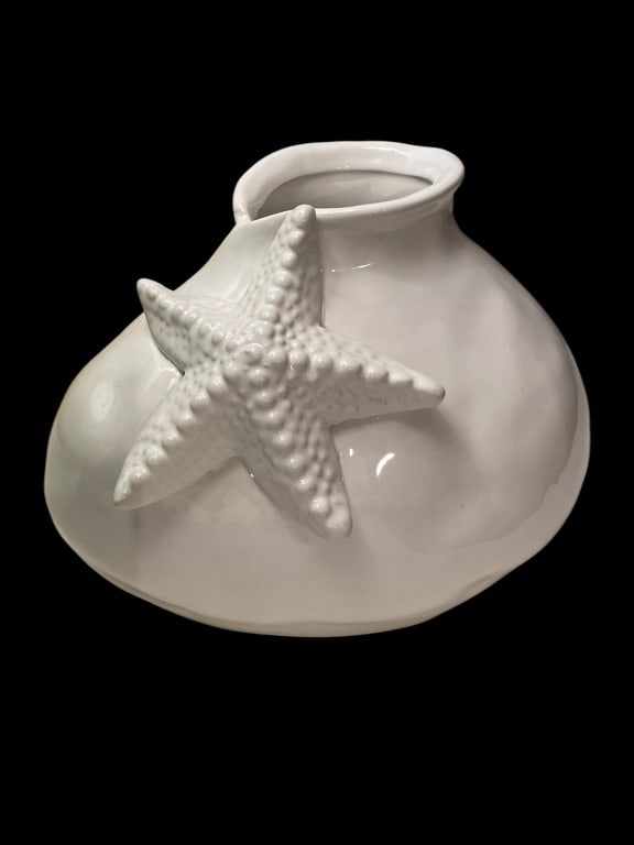WHITE CERAMIC VASE WITH STARFISH ACCENT 8": Glossy white ceramic vase featuring a prominent starfish motif on the front. Measures approximately 6 inches tall and 8 inches wide. The vase has a coastal, nautical charm, perfect for décor or flora