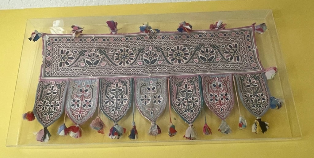 VINTAGE EMBROIDERED TEXTILE PANEL IN FRAME 46": Colorful embroidered textile panel featuring intricate floral patterns and decorative tassels. Displayed in a protective acrylic frame measuring approximately 22 x 46 inches. Excellent for wall decor,