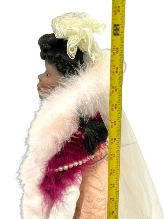 PORCELAIN DOLL WITH FEATHER STOLE AND VEIL: This porcelain doll stands approximately 18 inches tall and features dark curly hair, a lace and tulle veil, and a pink and magenta feather stole with faux pearl accents. The doll is dressed in period