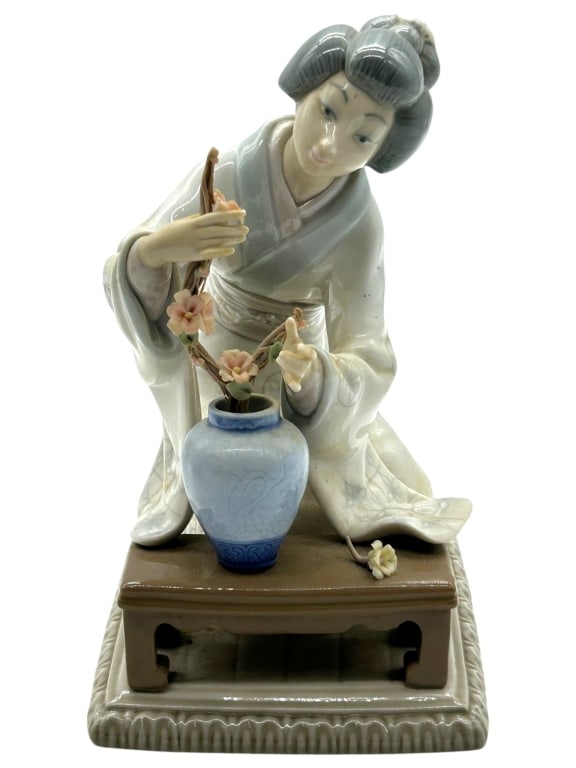 LLADRO GEISHA IKEBANA FIGURINE ON BASE 7.5": Elegant Lladro porcelain figurine depicting a seated geisha carefully arranging floral stems in a blue vase, rendered in the brand?s signature soft pastel palette with delicate hand-painted details.