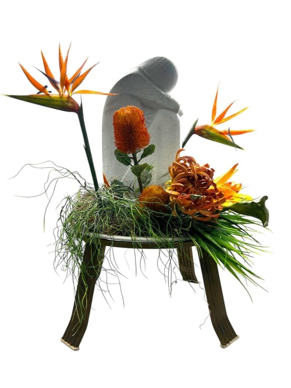 SCULPTED STONE FIGURE WITH FLORAL ARRANGEMENT 28": Sculpted stone figure in an abstract form, displayed on a stand. Features a floral arrangement with Bird of Paradise, Banksia, and assorted greenery. The arrangement is presented on a four-legged