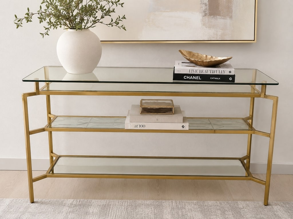 MODERN GLASS & METAL CONSOLE TABLE 55in: Modern console table featuring a brushed brass-finish metal frame with a clear tempered glass top, decorative inset glass center shelf, and mirrored lower shelf. The clean geometric silhouette and mix