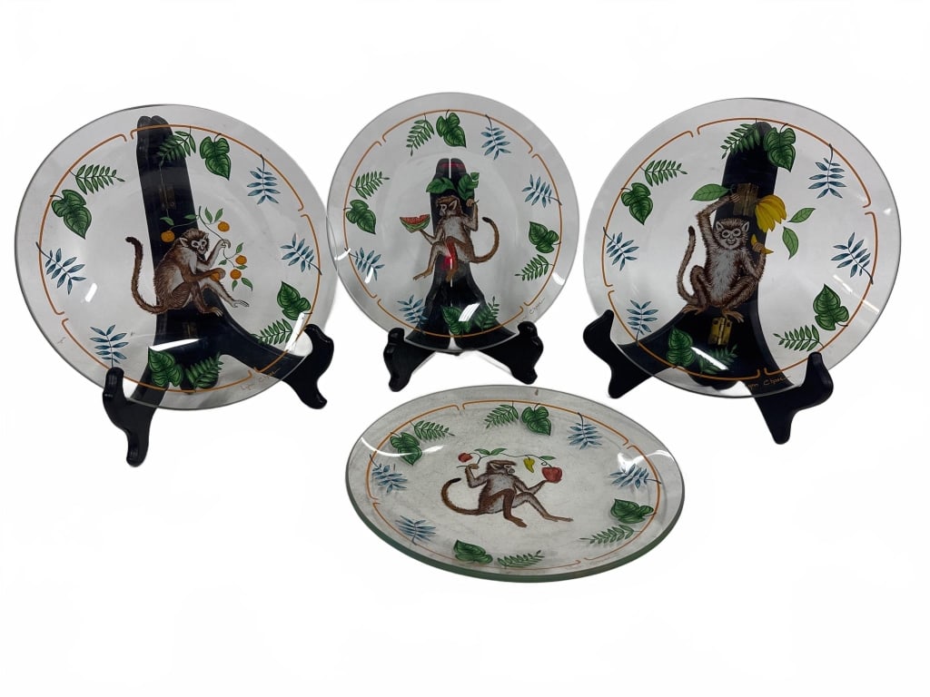 Lynn Chase Monkey Glass Plate Set: Four Lynn Chase glass plates, each featuring a whimsical monkey design with botanical accents. The plates are clear with multicolored painted details and each is signed by the artist. Plates measure a