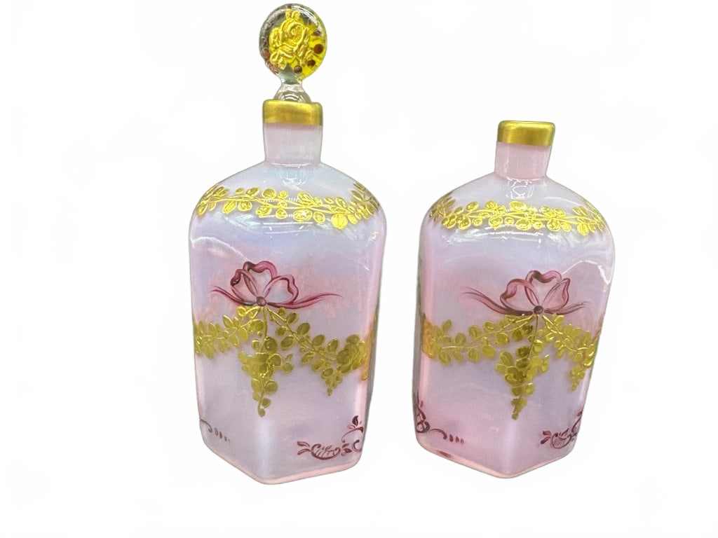 Pink Enameled Glass Decanters: Elegant pair of hexagonal pink glass decanters featuring hand-painted gold and rose floral enamel accents. One decanter includes a matching floral stopper. Both display gold trim at the neck. Approxim
