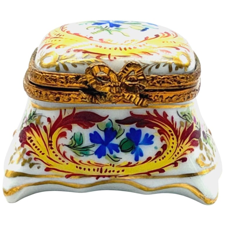 LIMOGES PEINT MAIN FLORAL BOX 1.5": Hand-painted Limoges porcelain trinket box featuring a central bouquet of pink roses framed by elegant gilt scrolling decoration. Finished with ornate brass-tone mounts and a decorative bow clasp, thi