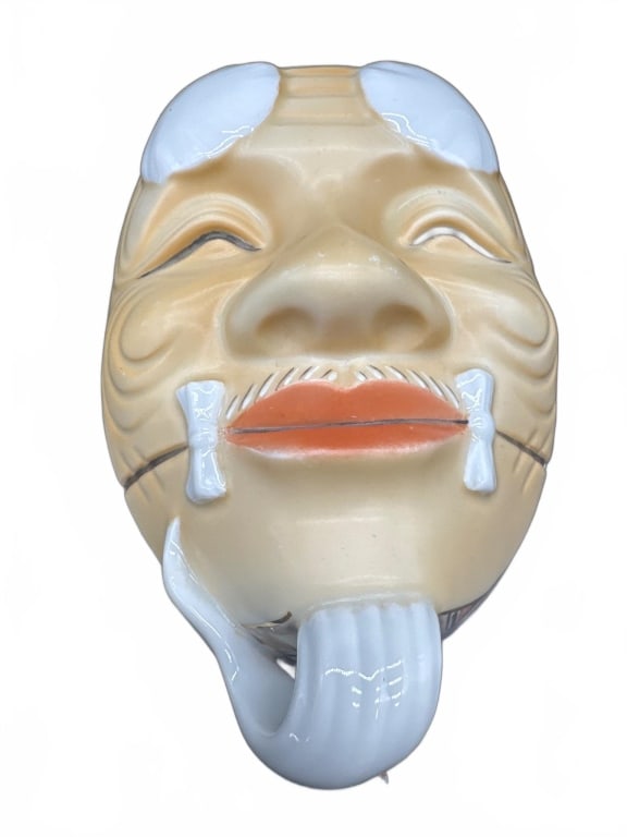 Porcelain Japanese Mask-Form Lidded Box: Porcelain lidded box shaped as a traditional Japanese mask, featuring a smiling face with raised features and painted details. The lid lifts to reveal a smooth white interior. The box measures approxi