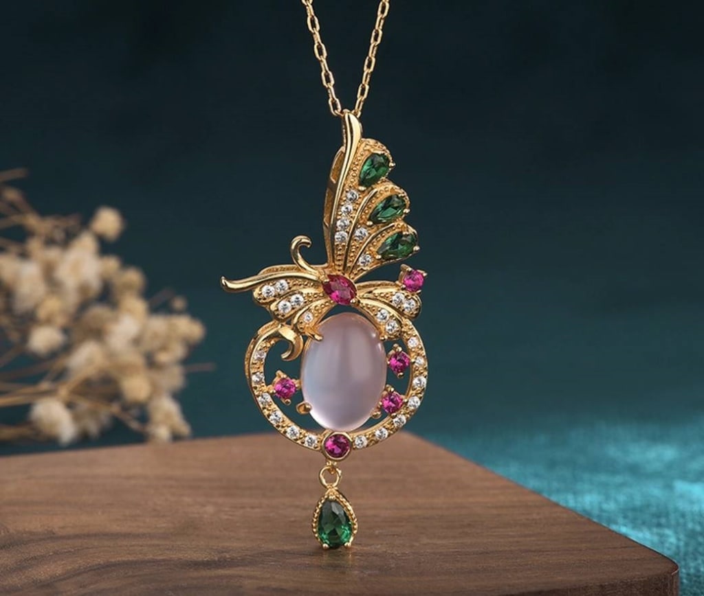 Crystal Butterfly Over Pink Jade 24K GP Necklace: 24K GP Gold necklace featuring a butterfly motif embellished with green and clear crystals, centered over an oval pink jade stone, with additional pink and green crystal accents and an ornate drop det
