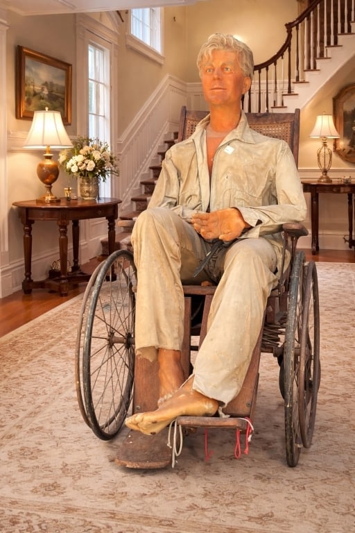 JOHN F KENNEDY WAX LIFE FIGURE ANTQ WHEELCHAIR: Life-size wax sculpture depicting President John F. Kennedy seated in an early-style wooden wheelchair, dressed in casual attire. The figure captures a rare and intimate portrayal, highlighting histor