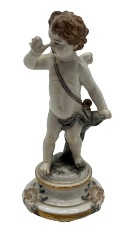 ANTIQUE GERMAN PORCELAIN PUTTO FIGURE 5.5": Antique German porcelain figurine depicting a standing putto or cherub modeled with curly brown hair, a draped garment and a small horn suspended from a blue and gilt shoulder strap. The figure stands