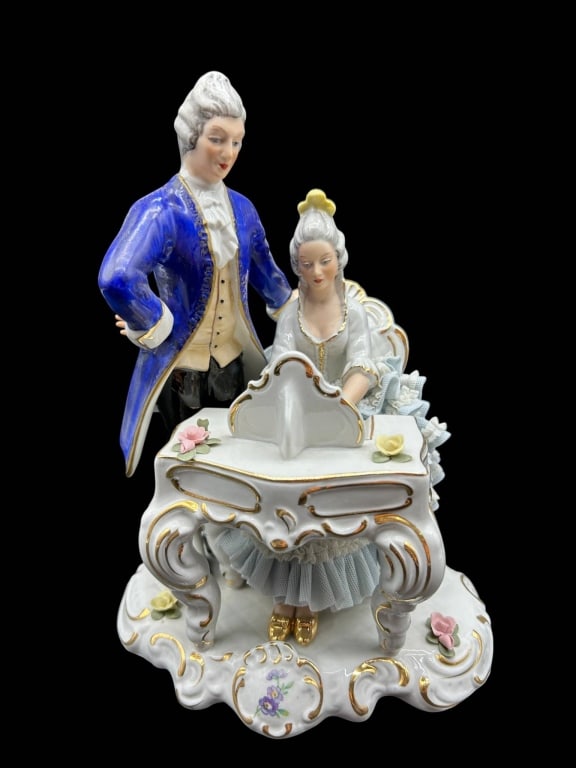 VINTAGE DRESDEN STYLE PORCELAIN PIANO COUPLE 9": Vintage Continental porcelain figural group in the Dresden tradition depicting an elegantly dressed 18th-century couple gathered at an ornate Rococo-style piano. The gentleman stands in a vivid
