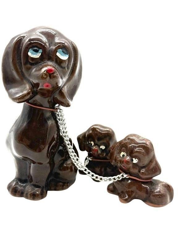 VINTAGE CERAMIC DOG FAMILY FIGURES 4": This charming vintage ceramic figurine set features a mother dog with two playful puppies connected by metal chain leashes. Finished in a glossy dark brown glaze with hand-painted facial details, the 