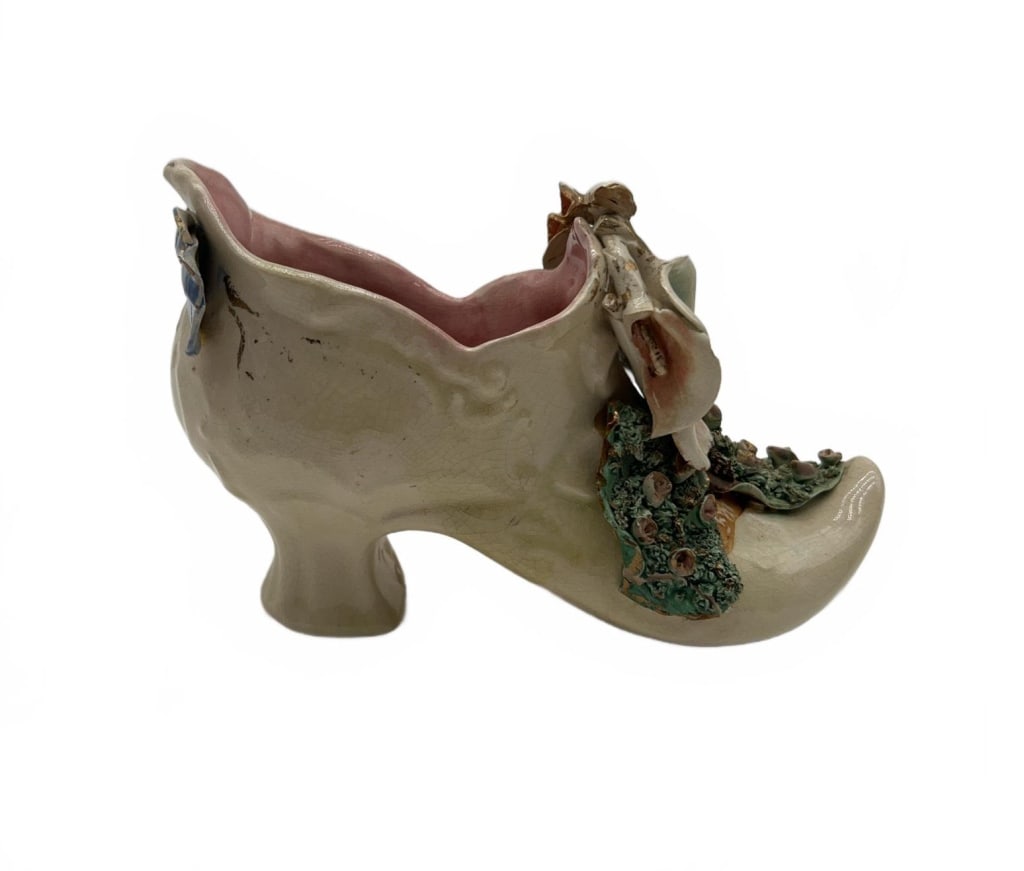 Porcelain Shoe Vase with Floral Decoration: Porcelain shoe-shaped vase featuring applied pastel flowers and gold accents, with a blue bow detail at the heel. Pink glazed interior. Measures approximately 6 inches long and 4.5 inches tall. No mak