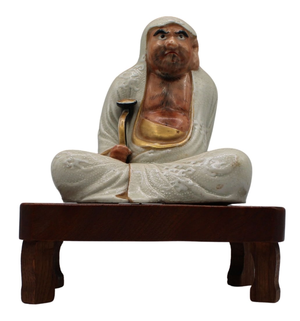 Japanese Taisho? Period Porcelain Damo Figure.: Japanese Taisho period porcelain figure of a Damo seated relaxed on a table. Small drill hole in the left ear. The Damo holds a black and gold staff and wears a white robe. The table is made of wood w
