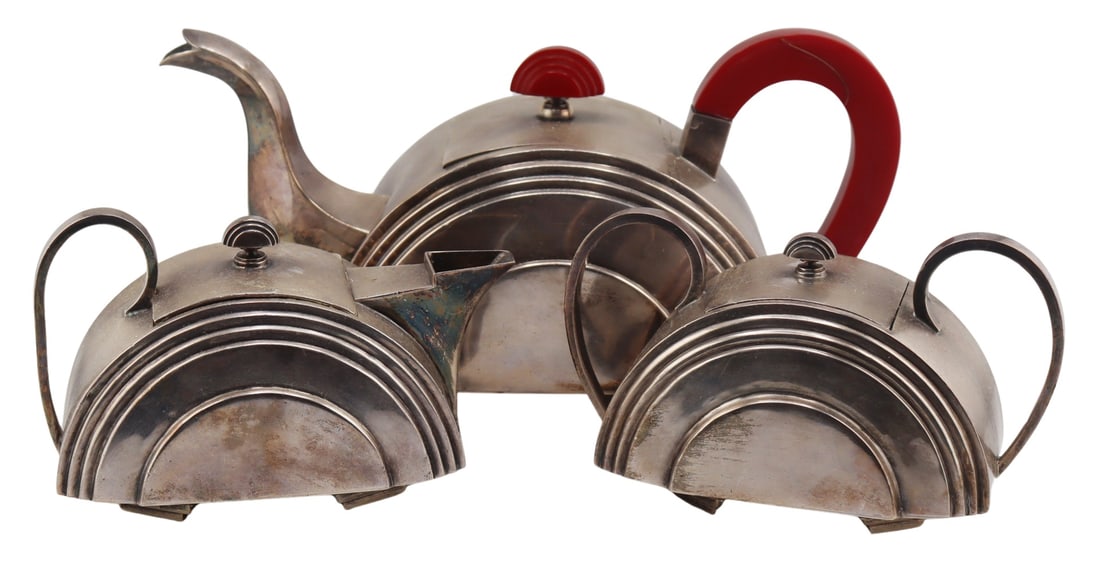 SILVER. 3 Pc. Art Deco Silver and Bakelite Tea Set: 3 pc. Art Deco silver tea service of half moon form with ribbed design including a teapot with carved cherry red Bakelite handle and finial, lidded creamer with spout, and lidded sugar. Stamped, "