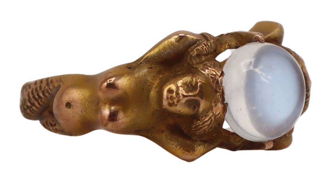 JEWELRY. Antique Gold and Moonstone Mermaid Ring.: Antique signed 11kt yellow gold ring in the form of two high relief mermaids displaying raised arms, exposed breasts, and woven textured tails. The mermaids' arms are raised and holding a prong-set ro