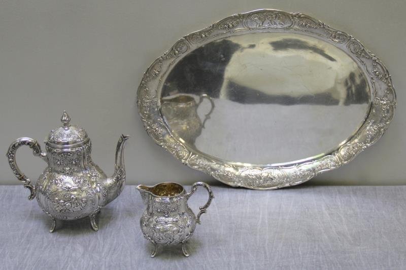 SILVER. German .800 Silver 3 Piece Tea Set.: Includes a footed teapot, footed creamer with gilded interior and oval serving tray - all with repousse floral swag and playing children motif. Total approx. weight is 79.79 troy oz. From a West 113th
