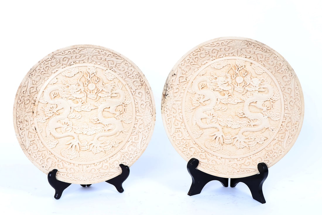 Pair of Cast White Dragon Plates: Lively pair of Chinese resin cast plates, imitating porcelain with pairs of dragons chasing flaming pearls. Each plate is heavily incised and detailed with floral and vines surrounding the rims,