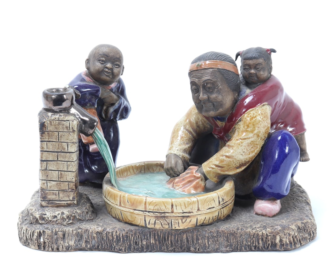 Chinese Porcelain Family, Grandmother & Grandchildren: Chinese porcelain statue depicting a grandmother and her grandchildren at a wash basin doing laundry. Granddaughter is climbing on grandma's back while the grandson pumps water into the basin. Size