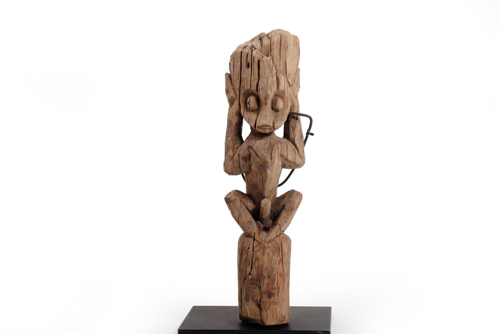 Borneo Carved Seated Male Figure: Borneo, 20th century or earlier. Carved wood figure depicting a seated male with hands raised beside the head, prominent eyes, and a columnar lower section. The attenuated torso, expressive posture,
