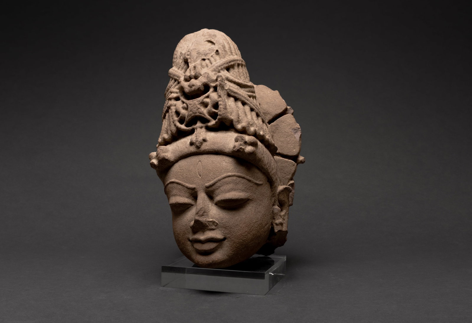 Indian Carved Stone Head of a Hindu Deity, 16th C.: India 16th century. A carved stone head of a Hindu deity Parvati, shown with a serene expression, almond shaped eyes, full lips, and a central forehead mark. The figure wears an elaborate tiered