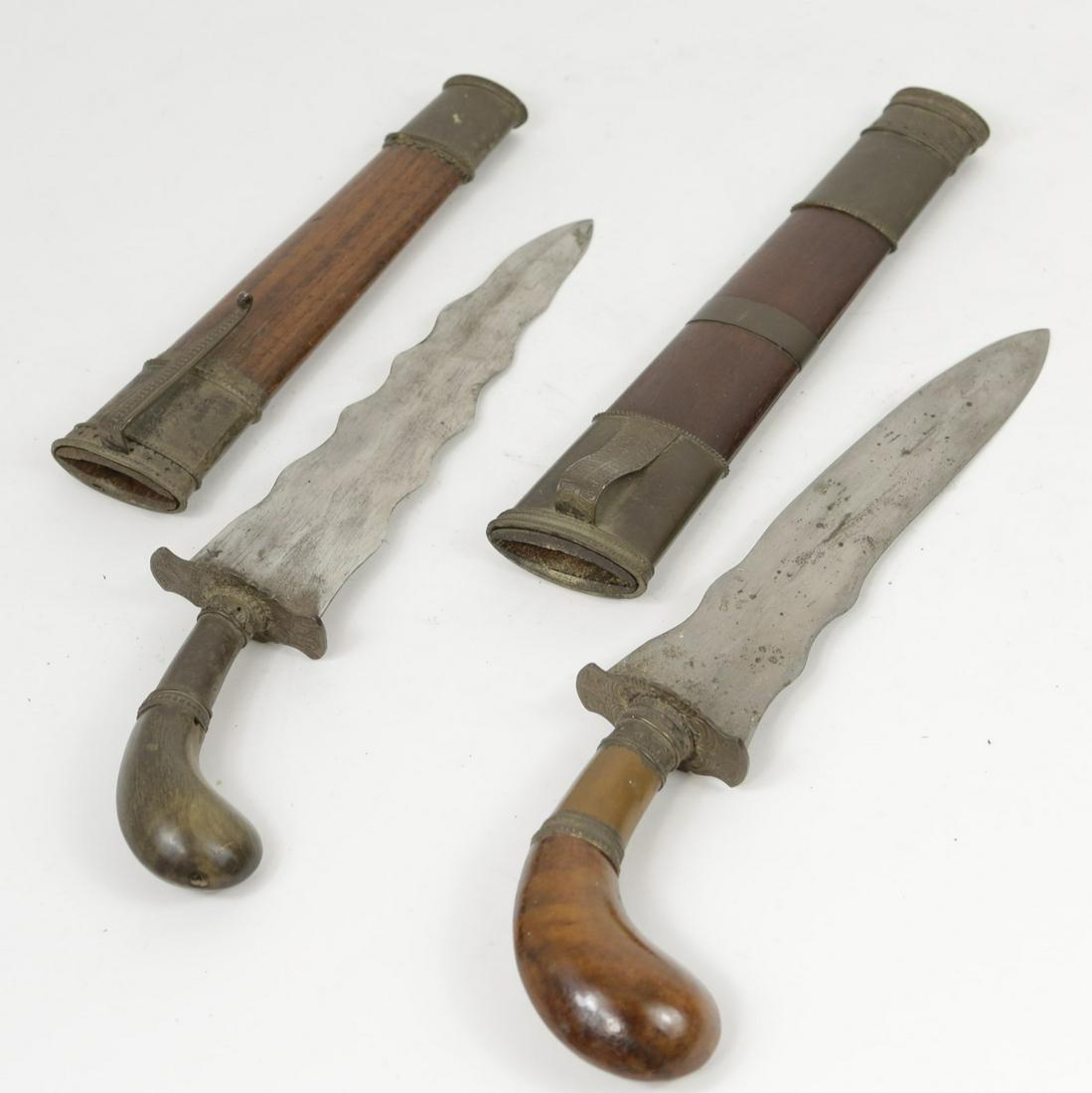 PR OF MORO OR KRIS DAGGERS w SHEATHS: Pair of Moro or Kris daggers with sheaths. Each having wooden pistol grip handle and scalloped blade. Wood handles and sheaths having metal mounts. Each measures 11 3/4" and 10 1/2" overall length.