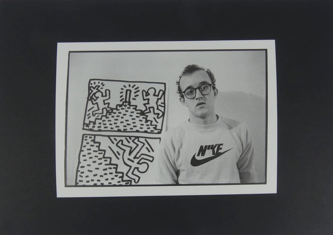 KEITH HARING PHOTO PRINT: Keith Haring photo print depicting the artist in Nike shirt with two works of art. Sheet measures 7 3/8" x 5 1/4". Mat measures 11 5/8" x 8 1/4".