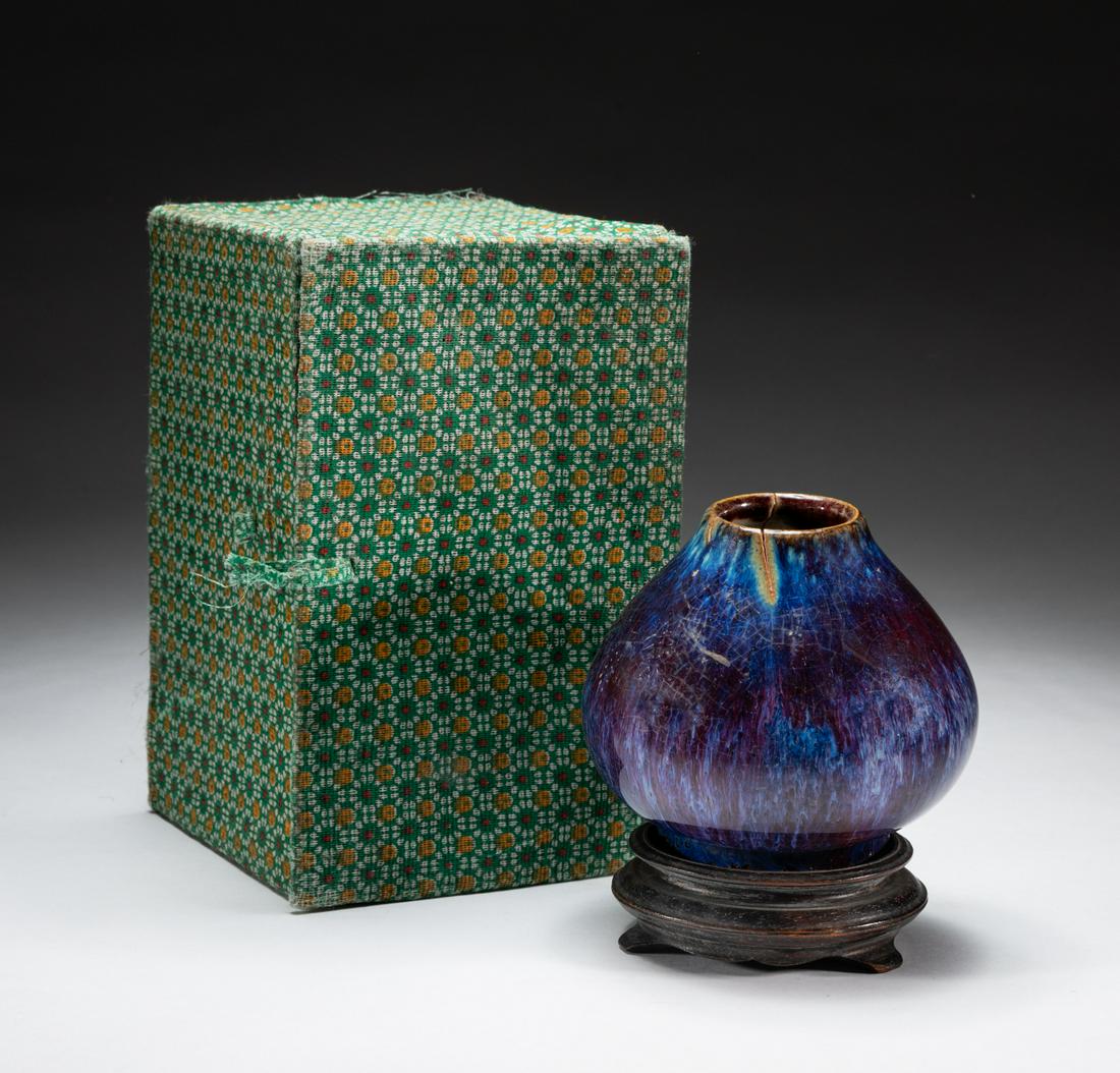 Chinese Flambe Glazed Porcelain Brush Pot: The robustly potted globular body rising from a recessed base to a narrow lipped mouth, of chicken heart type pot, applied with purplish glaze with raspberry streak, with a fitted wood stand, and box.