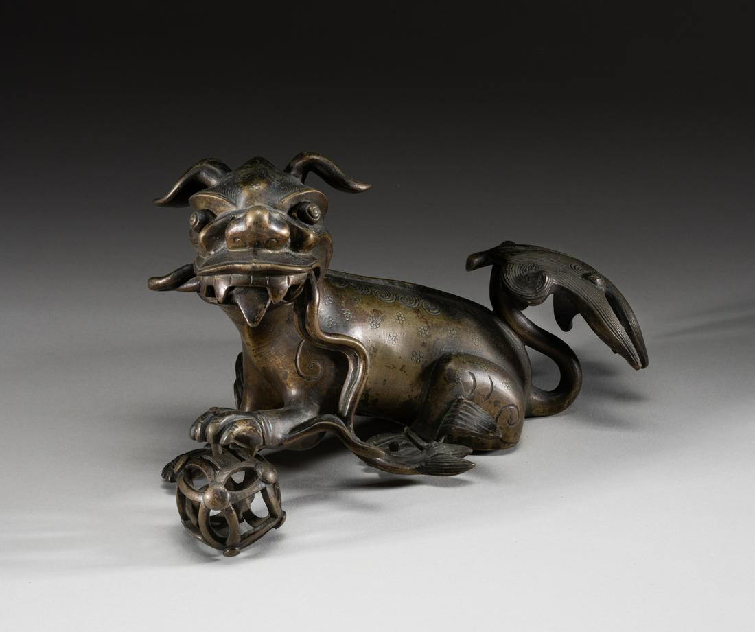 Chinese Bronze Foo Dog: Finley-casted bronze foo dog, the open mouth carrying a long ribbon, the front left paw on a hollowed ball, the body incised with curly fur and flower motifs in low relief, with detachable tail. 5.2in